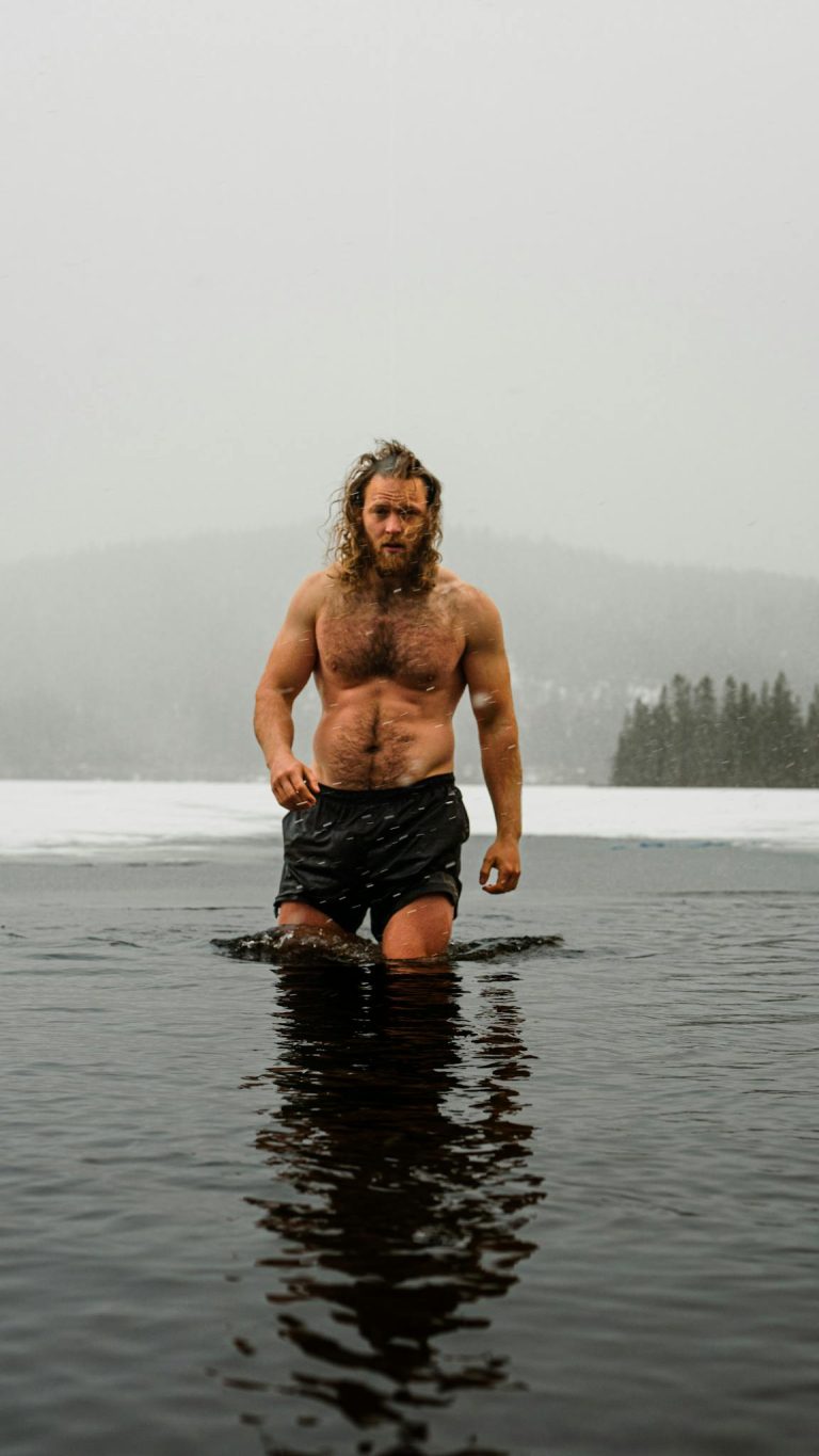 A topless bearded man braves icy lake water in a winter wilderness, embodying adventure.