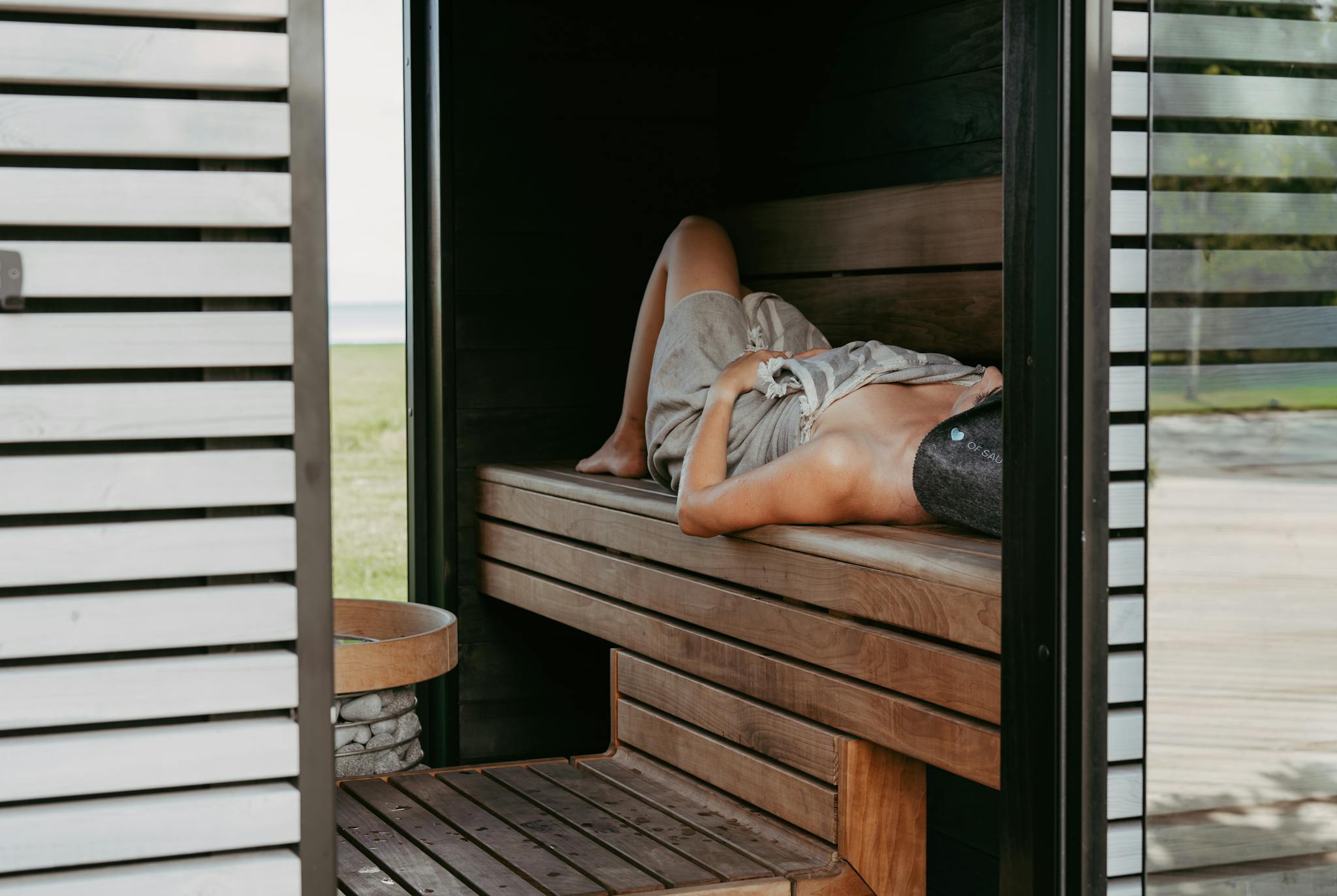 Person relaxing inside a modern sauna with Nordic design, lying with a towel and sauna hat for wellness.